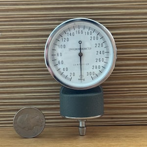 Puede incluir: Un esfigmomanómetro antiguo con una esfera blanca y una aguja negra, que muestra mediciones en mmHg. El medidor está montado sobre una base gris oscuro. Una moneda se coloca junto al medidor para la comparación de tamaños.