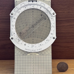 Pode incluir: Um computador de navegação Aristo-AviaT Nr. 613 vintage com uma régua de cálculo circular e uma grelha. O dispositivo tem uma moldura branca com marcações pretas. Uma moeda serve de escala.