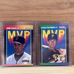 May include: Two baseball trading cards in protective cases. One features Will Clark of the Giants, the other Andy Van Slyke of the Pirates. Both cards have the word "MVP" prominently displayed.