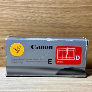 May include: A gray Canon focusing screen box with a yellow bird logo, red grid, and the text "Focusing Screen, Pantalla de enfoque, Verre de visee, Einstellscheibe, Schermo di messa a fuoco, E, C26-0556, D".