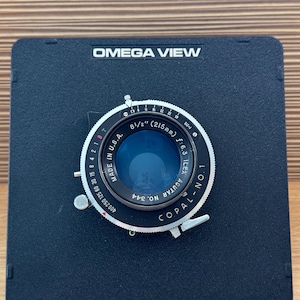 Può includere: Obiettivo per fotocamera Omega View nero con la scritta "8 1/2" (215mm) f:6.3 ILEX AGUTAR" e "MADE IN U.S.A.". L'obiettivo è montato su una base quadrata nera. L'obiettivo ha un anello argentato con indicazioni.