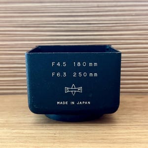 May include: A black camera lens hood with the text "F4.5 180 mm" and "F6.3 250 mm". The hood has a square shape and a circular base. The text "MADE IN JAPAN" is also visible.