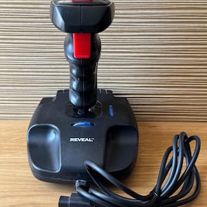 May include: Black joystick with a red button on top and a red button on the front. The base of the joystick has the word "REVEAL" printed on it. A black cable and connector are attached to the joystick.
