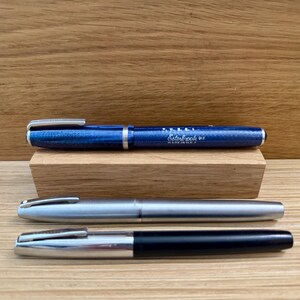 May include: Three fountain pens are displayed on a wooden surface. The top pen is blue with silver accents and the text "Esterbrook". The middle pen is silver, and the bottom pen is black and silver.
