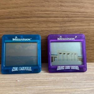 Op de afbeelding: Twee blauwe en paarse handheld elektronische spellen met het Intellivision-logo en de speltitels "Sea Battle" en "Black Belt Battle".