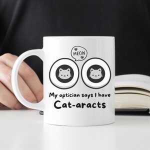 Może przedstawiać: Biały ceramiczny kubek do kawy z czarno-szarym rysunkiem dwóch oczu z kotami w środku. Tekst "MEOW" znajduje się w dymku nad oczami. Tekst pod oczami brzmi "Mój okulista mówi, że mam kocie zaćmy".