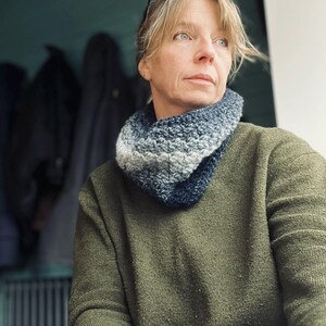 May include: A handmade, blue and gray crocheted cowl scarf. The scarf is worn around the neck and is made of a soft, textured yarn. The person in the image is wearing a green sweater and blue jeans.