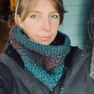 May include: A teal, purple, and blue crocheted cowl scarf. The scarf is worn over a dark gray jacket. The cowl is a thick, textured knit, with a triangular shape. The colors blend together in a gradient effect.