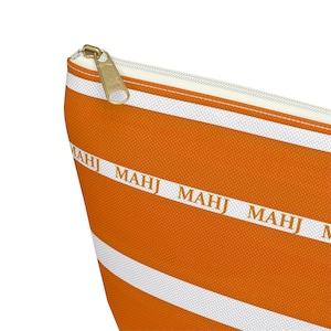 May include: Orange and white striped zippered pouch with the word "MAHJ" printed repeatedly. The pouch has a gold-colored zipper and a white interior. The design is a repeating pattern of orange stripes with white stripes.