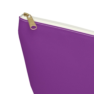May include: A close-up of a purple canvas pouch with a gold-colored zipper and a white zipper pull. The pouch is made of a textured fabric. The zipper is open.