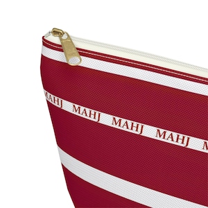 May include: A red and white striped cosmetic bag with a gold zipper. The bag features the word "MAHJ" printed in white along a white stripe. The bag is made of a textured material.