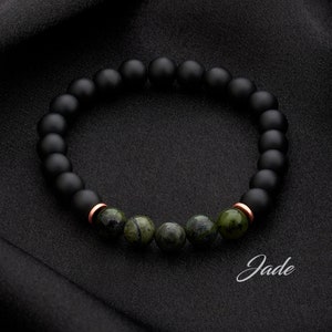 May include: A black beaded bracelet with green jade stones and copper accents. The bracelet is on a black fabric background. The word "Jade" is written in cursive below the bracelet.