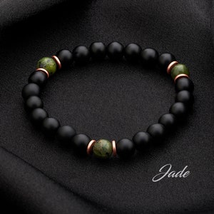 May include: A black beaded bracelet with three green jade beads and copper accents. The bracelet is on a black fabric background. The word "Jade" is written in cursive below the bracelet.