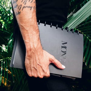 May include: A black planner with a silver spiral binding and the word "NOW" printed in white on the cover. The planner is being held by a person's hand.