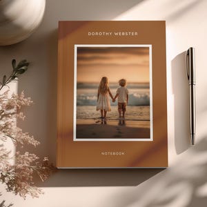 Puede incluir: Un cuaderno marrón con el texto "DOROTHY WEBSTER" en la parte superior y "NOTEBOOK" en la parte inferior. El cuaderno presenta una fotografía enmarcada de dos niños tomados de la mano en una playa. Un bolígrafo plateado descansa a la derecha.