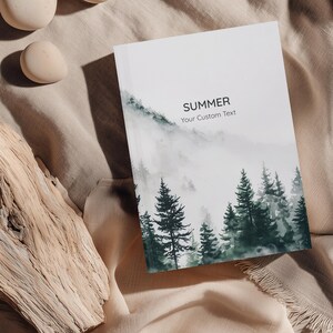 May include: A white journal with a watercolor forest scene on the cover, the word "SUMMER" and "Your Custom Text" are printed on the cover. The journal is on a beige cloth with decorative stones and wood.
