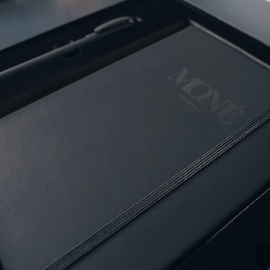 May include: A black leather-bound journal with the word "MON&Eacute;" and the text "journal" embossed on the cover. The journal is in a black box with a black pen.