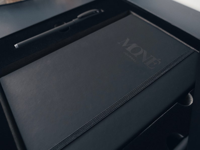May include: A black leather-bound journal with the word "MON&Eacute;" and the text "journal" embossed on the cover. The journal is in a black box with a black pen.