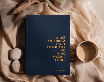 Peut inclure: Un journal bleu marine avec des lettres dorées qui dit "A List of Things I Will Eventually Do In No Specific Order" par Megan. Une tasse de café et des pierres décoratives sont à côté.