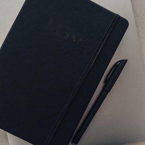 May include: A black notebook with the word "MOVE" embossed on the cover, a black pen is lying on top of the notebook.