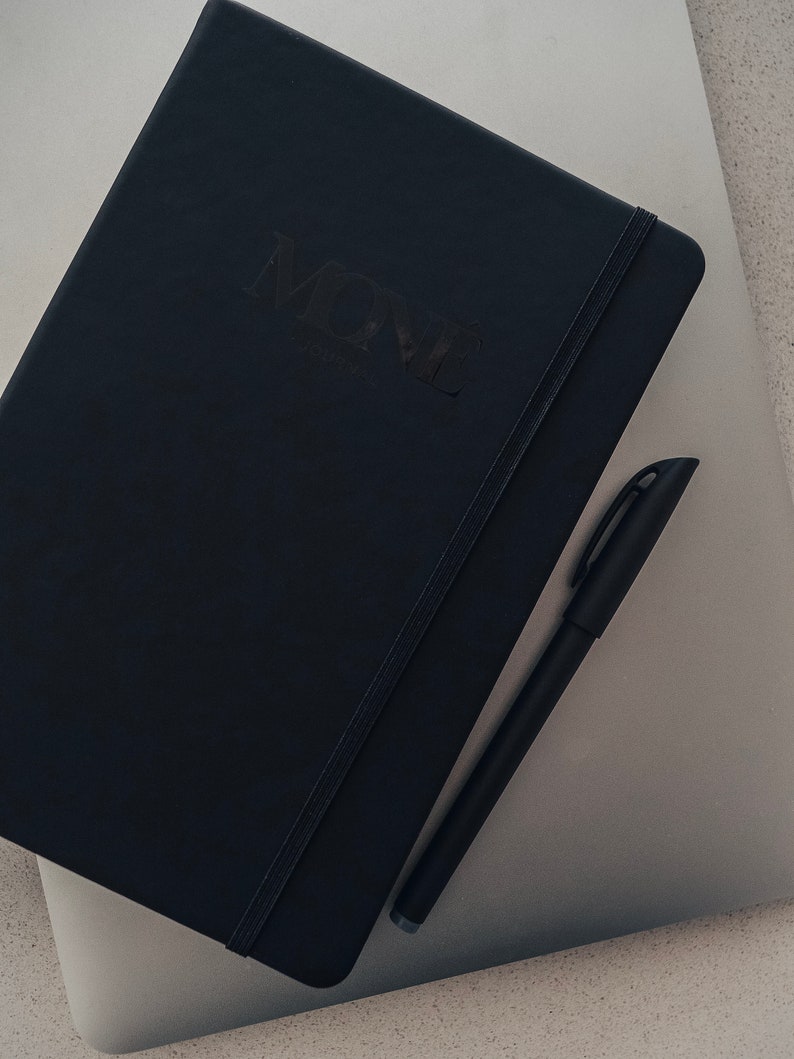 May include: A black notebook with the word "MOVE" embossed on the cover, a black pen is lying on top of the notebook.