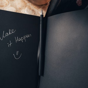 May include: A black notebook with a silver pen lying open on a table. The notebook has the words "Make it Happen" written on the left page in white ink, along with a smiley face.
