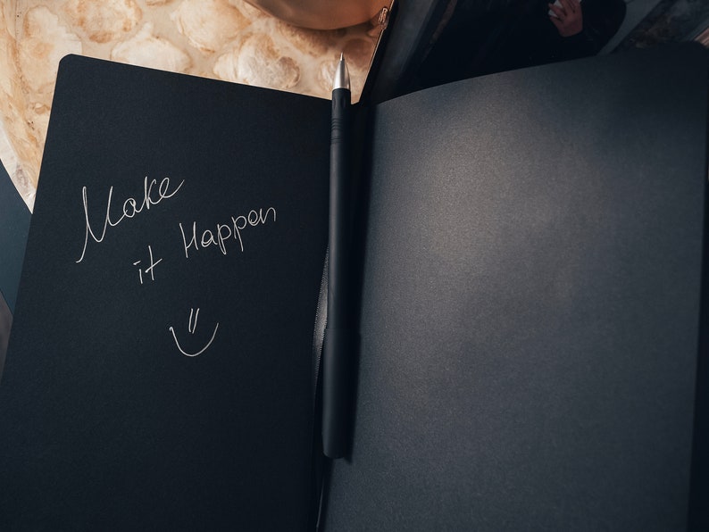 May include: A black notebook with a silver pen lying open on a table. The notebook has the words "Make it Happen" written on the left page in white ink, along with a smiley face.