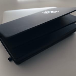 May include: A black leather-bound notebook with a white inscription on the cover. The notebook is closed and lying on a white surface.