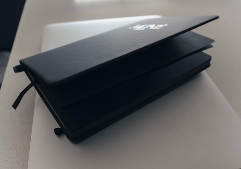 May include: A black leather-bound notebook with a white inscription on the cover. The notebook is closed and lying on a white surface.