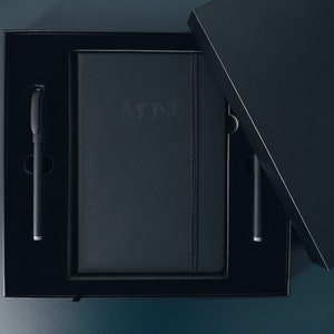 May include: A black box with a black notebook and two black pens inside. The notebook has the word "MONE" embossed on the front.