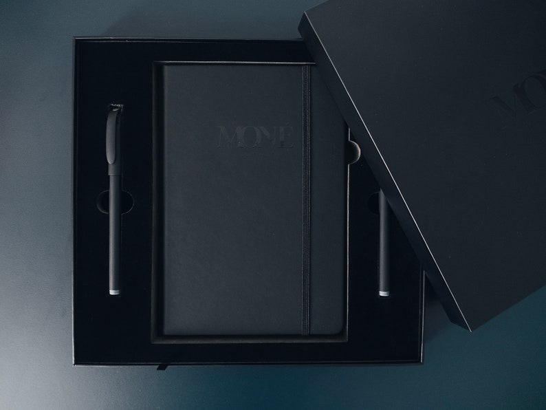 May include: A black box with a black notebook and two black pens inside. The notebook has the word "MONE" embossed on the front.