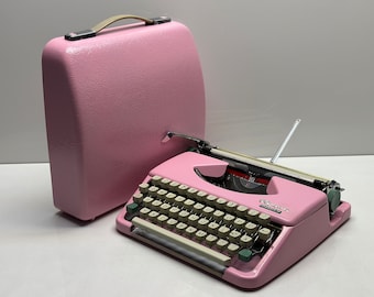 Puede incluir: Una máquina de escribir vintage rosa con una funda de transporte rosa a juego. La máquina de escribir tiene un teclado de color crema con teclas verdes y una palanca de retorno de carro plateada.