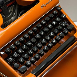 Puede incluir: Primer plano de una máquina de escribir Olympia naranja con teclas negras. Las teclas están dispuestas en un diseño QWERTY, con letras, números y símbolos. La palabra "Olympia" es visible.