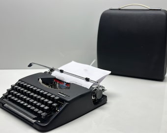 May include: A black vintage typewriter with a white paper insert and a black carrying case. The typewriter has a standard keyboard layout with black keys and a silver carriage return lever.