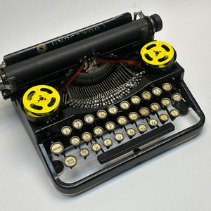 May include: A black vintage Underwood typewriter with yellow spools of ribbon. The typewriter has a standard keyboard layout with keys for letters, numbers, and punctuation.