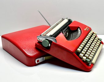 Puede incluir: Una máquina de escribir roja con un teclado blanco y verde, en una maleta de transporte roja. La máquina de escribir tiene una palanca de retorno de carro plateada y una palanca de liberación de papel plateada.