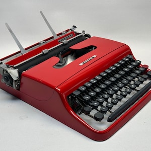 Könnte beinhalten: Eine rote Vintage-Schreibmaschine mit schwarzen Tasten und einem silbernen Wagenrücklaufhebel. Die Schreibmaschine trägt ein Etikett mit der Aufschrift "Lettera 22".