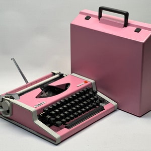 May include: A pink vintage Olympia portable typewriter with a matching pink carrying case. The typewriter has a black keyboard and a white carriage return lever.
