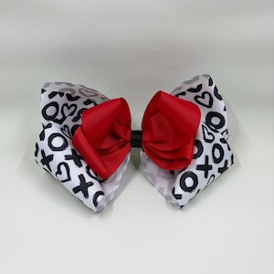 May include: A handmade hair bow featuring a red center and white and black patterned wings. The wings are decorated with hearts, X's, and O's. The bow is a perfect accessory for Valentine's Day or any special occasion.