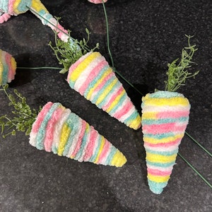May include: Four colourful striped carrot-shaped decorations with green stems. The carrots are made of fabric and have a fuzzy texture. They are arranged on a black surface.