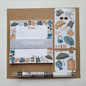 May include: A travel-themed notepad set with a pen. The notepad features illustrations of travel items like a camera, airplane, and coffee cup. A bookmark with "Bon Voyage" text and travel stickers are included. The pen has a white barrel and a decorative design.