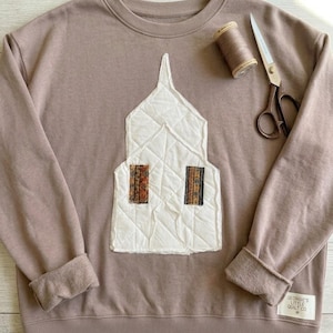 May include: A taupe crewneck sweatshirt featuring a white quilted church-shaped applique with two small patterned fabric windows. A spool of matching thread and metal scissors rest on the fabric, with a small label reading Georgie's Little Quilt Co.