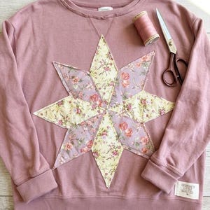 May include: A dusty rose long-sleeve sweatshirt featuring a large eight-pointed star applique made of floral fabric in shades of yellow and pink. A spool of matching thread and metal scissors lie nearby. A label reads Georgie's Little Quilt Co.