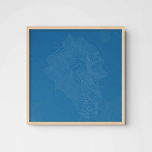 May include: A square framed wall art piece featuring a blue background with white topographic contour lines depicting a mountain or landform. The frame is made of light-colored wood, providing a clean border around the blue map illustration.