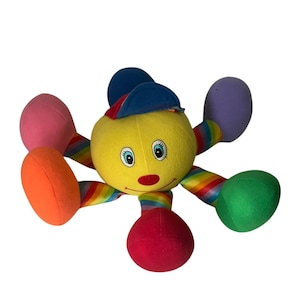 May include: A plush toy featuring a round yellow head with a smiling face, blue eyes, and a red nose, wearing a blue baseball cap. The toy has six multi-colored limbs with rainbow-striped segments and solid-colored ends in pink, orange, red, green, and purple.