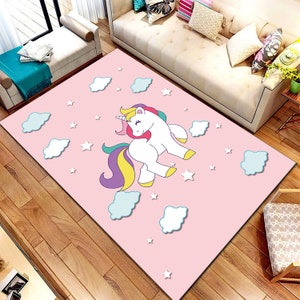 Può includere: Un tappeto rosa con un unicorno bianco con una criniera e una coda arcobaleno. L'unicorno è circondato da stelle bianche e nuvole blu.