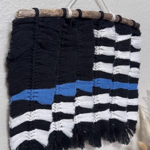 May include: A wall hanging made of black, white, and blue macrame cord knotted onto a natural wooden branch. The design features a solid black section on the left and alternating black and white horizontal stripes on the right, with a single blue stripe.