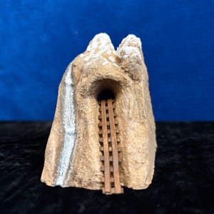 May include: A miniature mountain model with a tunnel entrance and a wooden ladder. The mountain is primarily tan with white accents on the peaks and a silver waterfall effect. The tunnel is dark, and the ladder is brown.
