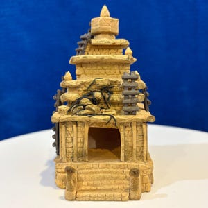 Può includere: Un tempio in miniatura color beige con un design a più livelli. La struttura presenta un'apertura centrale, gradini in pietra e piccole scale sui lati. Il tempio è adornato con accenti scuri e usurati, che suggeriscono età e usura.