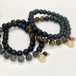 Könnte beinhalten: Drei Perlenarmbänder mit goldenen Akzenten. Die Armbänder bestehen aus schwarzen Onyx-, Tigerauge- und Rauchquarzperlen.